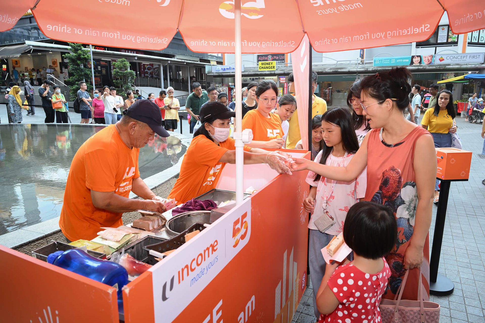 People, including children, receive food from vendors at an outdoor "Income" branded stall under umbrellas.