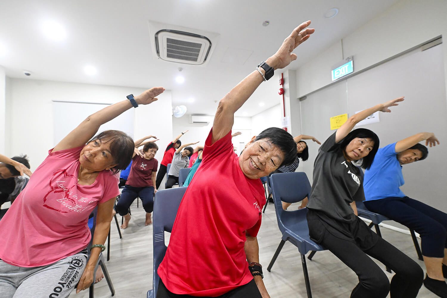Smiling seniors stretching in chairs, raising arms to the side.