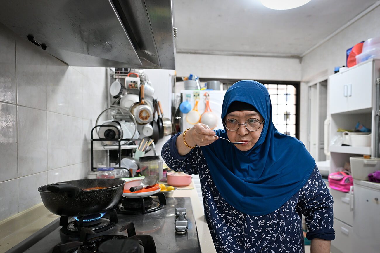 Elderly woman in a blue hijab tastes food from a spoon in a kitchen.