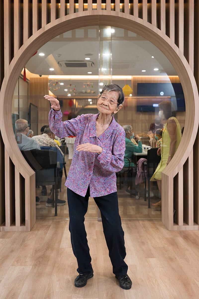 Elderly Asian woman joyfully dancing, framed by a decorative wooden circle.